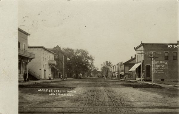 View down unpaved street in a small town lined with businesses. A sign painted on the side of a building on the right reads: "Monona House." Caption reads: "Main St. Looking North, Lone Rock, Wis."