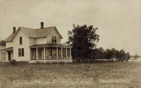 View across lawn towards a two-story dwelling next to a lake. Three people are sitting in the screened-in porch. Caption reads: "A.J. Larson's Residence, Long Lake, Wis."