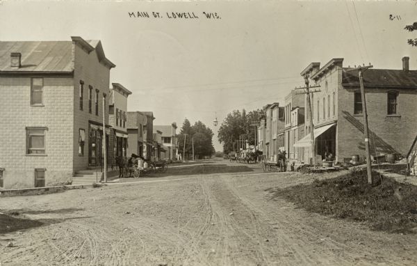 View of a small town street lined with businesses. Horses and carts with milk cans are in the street. Caption reads: "Main St., Lowell, Wis."