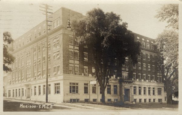 View across street towards a five-story corner building with entrances at the front and the side. An electric YMCA sign is near a tree in front. Caption reads: "Madison - Y.M.C.A."