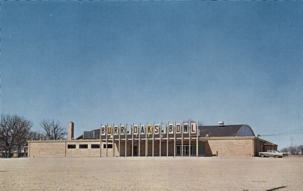 View across parking lot towards the bowling alley located at 2202 South Park Street. There is a large electric sign above the entrance. Text on reverse reads: "The most complete bowling facilities in Central Wisconsin. 32 automatic Brunswick Crown Imperial Lanes, beautiful cocktail lounge, and restaurant. Plenty of Parking Space."