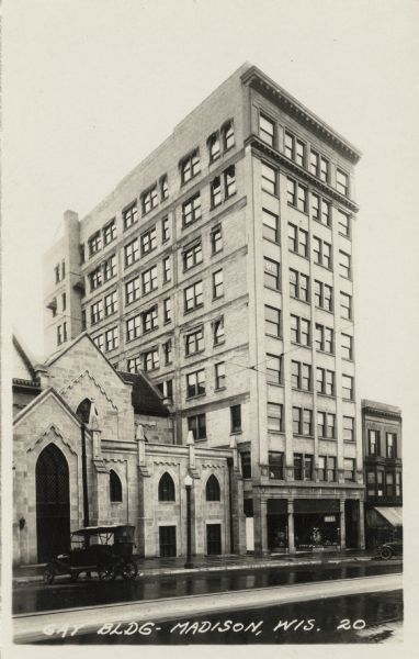 View across N. Carroll Street towards the Gay Building (now the Churchill Building), Madison's first skyscraper. A nine-story building located at 16 North Carroll Street. There is a sign for "Metropolitan Life" in the sixth floor windows. A pharmacy is on the ground floor, and automobiles are parked at the curb. Grace Episcopal Church is next door on the left. Caption reads: "Gay BLDG - Madison, Wis."