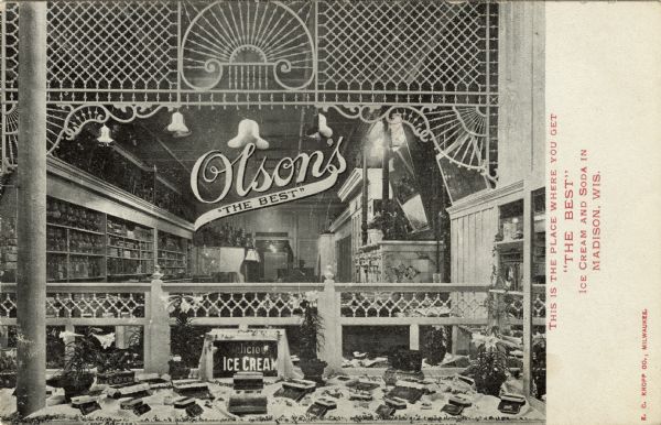View of the window display of an ice cream parlor. There is a decorative wood screen hanging above a display of boxes of chocolate and signs for ice cream in the foreground. Inside the store are glass jars on shelves behind a counter on the left. Text on front reads: "This is the place where you get 'The Best' ice cream and soda in Madison, Wis."