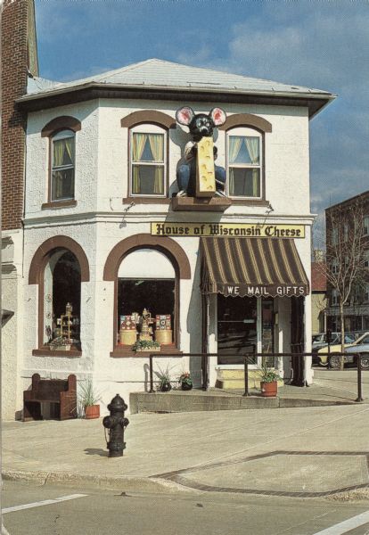 Exterior view of a cheese shop with a sculpture of a mouse eating cheese above the entrance located at 101 N. Hamilton Street. Cheese is on display in the arched windows near the entrance. The sign on the awning above the doorway reads: "We Mail Gifts." Text on back reads: "The Cheesehouse with the Mouse, Located in the shadow of the state capitol, we've offered the finest cheese products from America's Dairyland for over 35 years. A small business filled with lots of Wisconsin Pride."