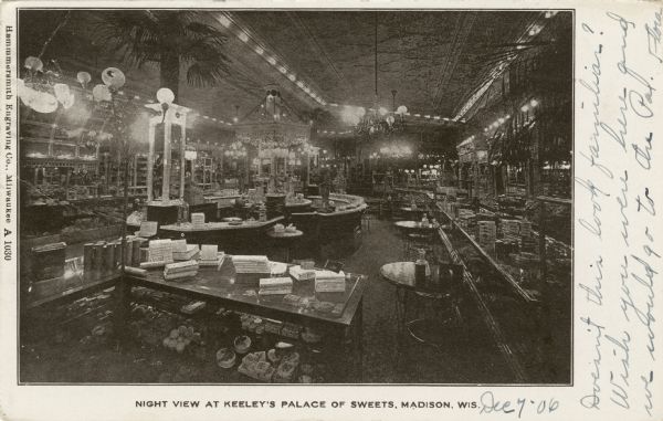 Elevated view of a sweet shop and soda fountain located at 112 State Street. In the foreground is a glass counter, and behind it on the left is the soda fountain, with seating along a long glass counter on the right. The space also includes a tin ceiling, mirrors along the walls, and decorative light fixtures. Caption reads: "Night View at Keeley's Palace of Sweets, Madison, Wis."