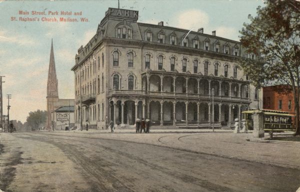 View of the Park Hotel, located on the corner of South Carroll Street and West Main Street. The church is further down the block. There is a sign on the building behind the hotel that reads: "The New St. Nicholas Restaurant." A streetcar is coming around the square on Carroll Street. Pedestrians are in the street and on the sidewalks. Caption reads: "Main Street, Park Hotel and St. Raphael's Church, Madison, Wis."