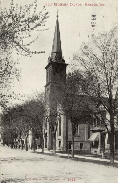 View down street towards the Catholic church located on West Johnson Street. There is a clock tower on the steeple. Caption Reads: "Holy Redeemer Church, Madison, Wis."