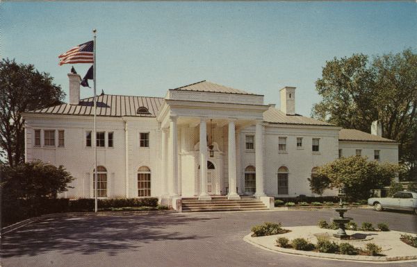 Exterior view from driveway of the Governor's Mansion in Maple Bluff. Flags are flying from a flagpole in the yard on the left. Columns frame the front entrance, and a fountain is in the foreground.
