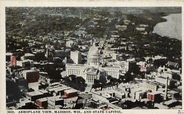 Aerial view of the Wisconsin State Capitol and the square from over King Street. Lake Mendota is on the right. The State Historical Society is at the end of State Street. There is a water tower on East Washington Avenue in the lower right corner. Caption reads: "Aeroplane View, Madison, Wis. and State Capitol."