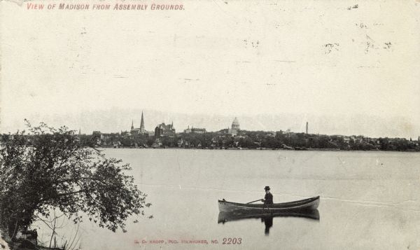 View from shoreline towards the Madison skyline from across Lake Monona. Landmarks include a church, the Park Hotel, Madison City Hall, the old Capitol and the East Washington Avenue water tower. A man is in a rowboat in the foreground. Caption reads: "View of Madison from Assembly Grounds."