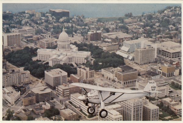 Aerial view of the Capitol facing Lake Mendota. The Spirit of St. Louis is superimposed on the postcard. Landmarks include the glass bank, the YWCA and James Madison Park.