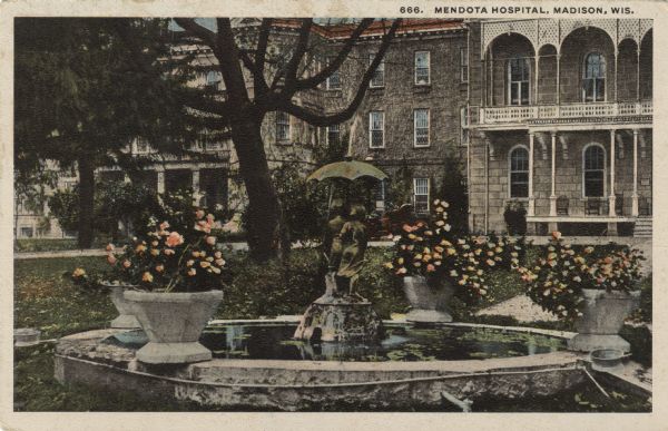 The front lawn of Mendota Hospital. In the fountain is a statue of children under an umbrella, with flowers in planters on the edge of the circular wall around the fountain. Caption reads: "Mendota Hospital, Madison, Wis."