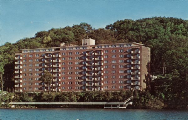 View across water towards the large building on the shoreline of the lake. Text on reverse reads: "The Commodore on Lake Mendota at 3100 Lake Mendota Drive. A retirement home with a nursing section. Wisconsin's largest and most luxurious. Gracious living at reasonable rates."