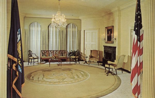 Interior view of the reception room at the Governor's Mansion. A U.S. Flag and a Wisconsin flag are flanking the doorway. The state seal of Wisconsin is on the circular rug. A portrait is hanging over the fireplace.