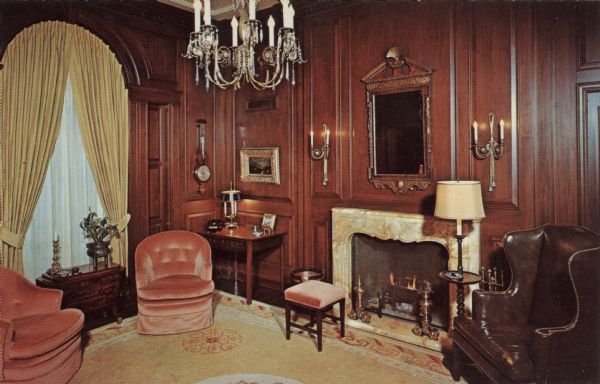 Interior view of the library at the Governor's mansion. The walls are covered with wood paneling, draperies are on an arched window on the left, and a mirror is hanging above the fireplace.