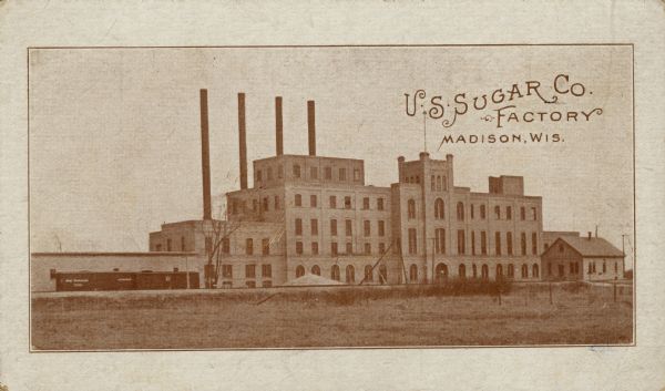 View across field towards a sugar factory, with four smokestacks in the background. Railroad cars are on railroad tracks along the front of the building.