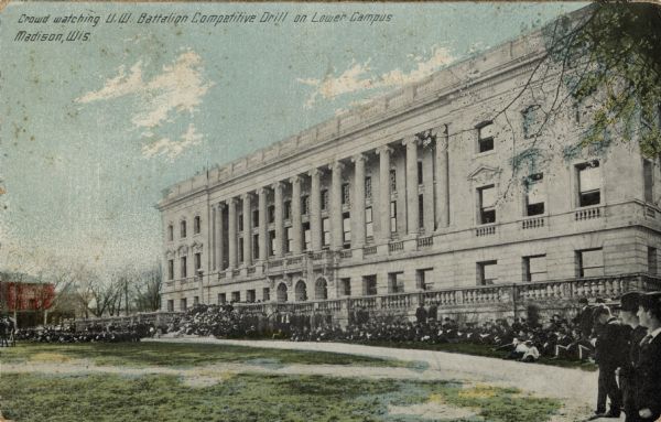 Hand-colored postcard of a crowd on and around the front steps of the State Historical Society building watching a battalion drill (out of the picture). Soldiers are on the left. Caption reads: "Crowd Watching U.W. Battalion Drill on Lower Campus, Madison, Wis."