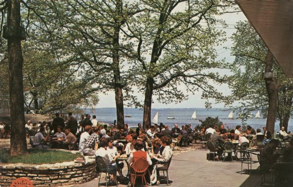 View of the Terrace at Memorial Union with students and other people sitting around tables in colorful Terrace chairs. Sailboats are out on the lake.