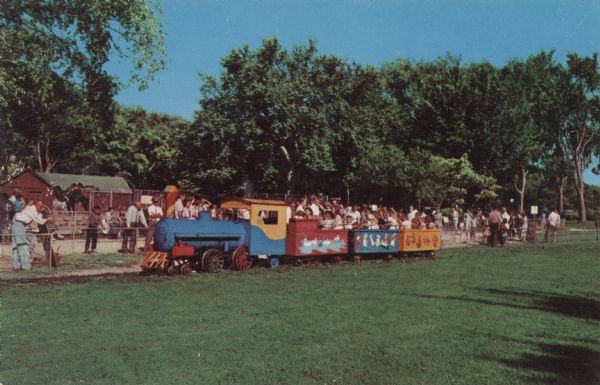 View of people riding the Kiddie Train at the Vilas Park Zoo. People behind the train are standing and watching the train near a fence, and there is a camel in an enclosure near buildings in the background.