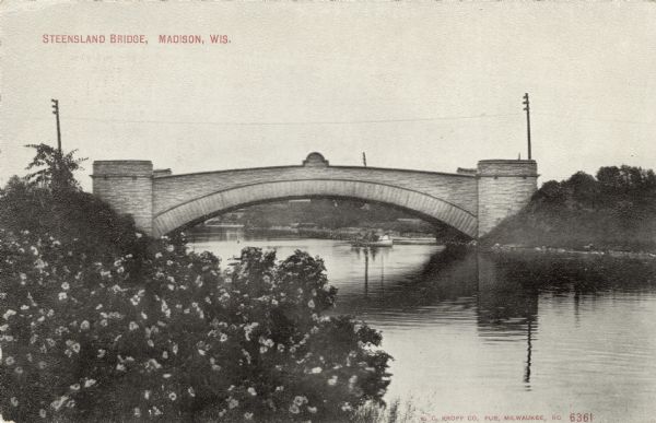 View from shoreline towards the Steensland Bridge crossing the Yahara River at Tenney Park. There are people in a boat just under the bridge. Caption reads: "Steensland Bridge, Madison, Wis."