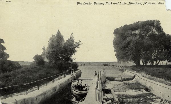 View of the locks at Tenney Park where Lake Mendota meets the Yahara River. A boat with five people is passing through the canal. Caption reads: "The Locks, Tenney Park and Lake Mendota, Madison, Wis."