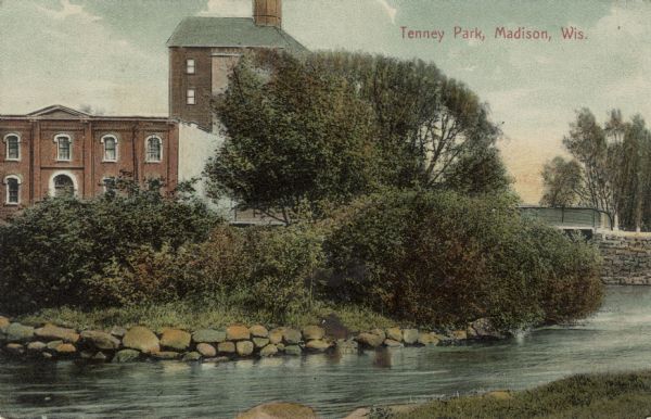 View from shoreline across river towards an industrial building near the Yahara River. A bridge is behind trees and bushes on the right. Caption reads: "Tenney Park, Madison, Wis."
