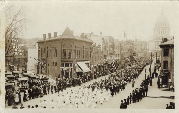 Elevated view of a patriotic parade on State Street, with marchers coming from the Capitol Square. The intersection of East Johnson and State Street is in the foreground. Flags are flying over the street and over the sidewalks. Several automobiles are on Johnson Street behind the line of spectators.