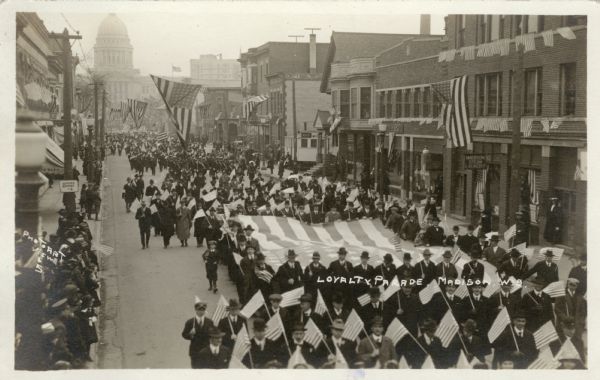 Elevated view of a patriotic parade on State Street, coming from the Capitol Square. Men in suits and overcoats walking down the street are carrying flags, and another group is carrying a large flag horizontally down the street. More flags are hanging over the street and sidewalks.
