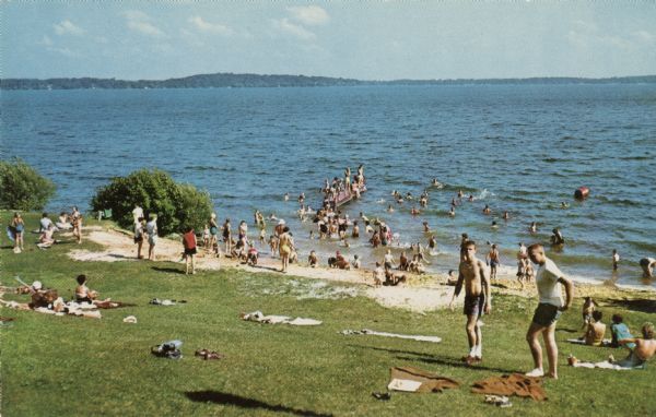 View of a crowded bathing beach on Lake Monona. Blankets are spread out on the lawn.