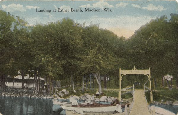 View of the dock at Esther Beach, Lake Monona. People are sitting in boats moored along the pier near the shoreline. Caption reads: "Landing at Esther Beach, Madison, Wis."
