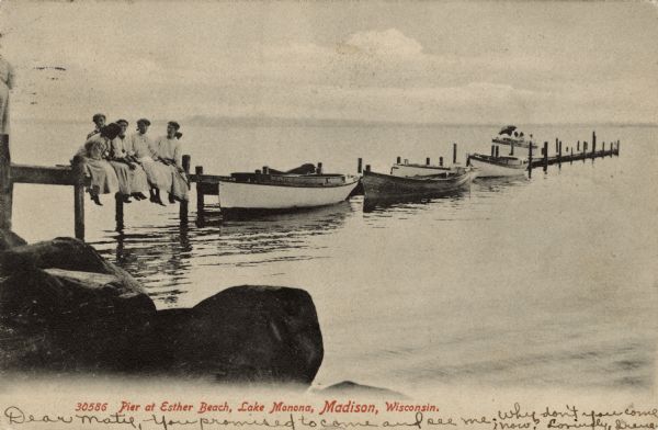 View from shoreline towards five women sitting together on the pier at Esther Beach. Boats are moored along the pier. People are in a boat near the end of the pier. Caption reads: "Pier at Esther Beach, Lake Monona, Madison, Wisconsin."
