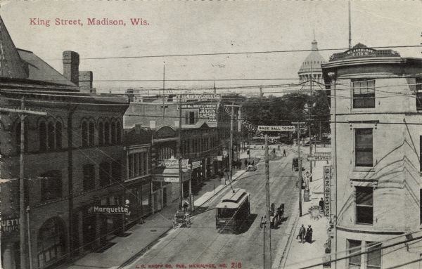 Elevated view of King Street looking towards the Capitol Square. There is a streetcar and a horse-drawn vehicle on the street. The Sherlock and Sons building is on the right. The Marquette Hotel and Majestic Theater is on the left. Caption reads: "King Street, Madison, Wis."