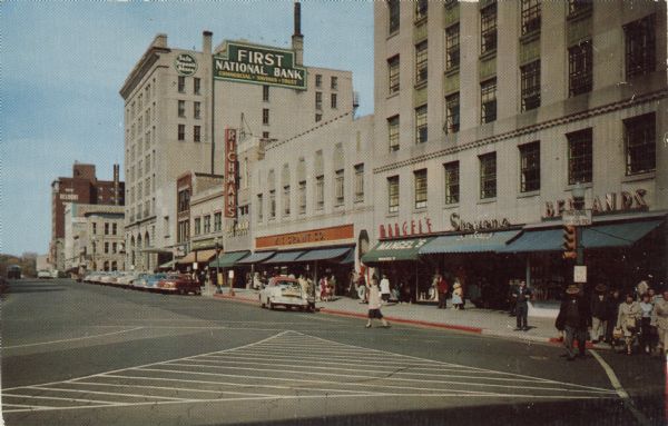 A view across Pinckney Street at the corner of Main Street. The two blocks along the Square include storefronts for Stevens, Mangel's, and Richman's. The First National Bank is on the corner at East Washington Avenue. The Belmont Hotel further down the street is at the corner with Mifflin Street.