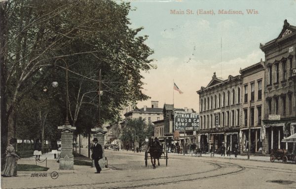 View of Main Street along the Capitol Square facing east. Streetcar tracks in the street. Businesses include a piano store, a dry goods store and a restaurant. Caption reads: "Main St. (East) Madison, Wis."
