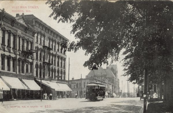 View of Main Street along the Capitol Square near the intersection with S. Carroll and S. Hamilton Streets facing west. A streetcar is coming up the street. Caption reads: "West Main Street, Madison, Wis."