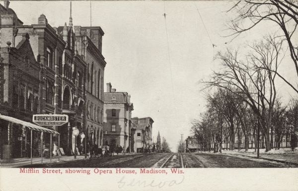 View down Mifflin Street, facing east along the Capitol Square. The Opera House and City Hall are on the left. There is a large sign over the sidewalk for Buckmaster Jeweler. Caption reads: "Mifflin Street, showing Opera House, Madison, Wis."