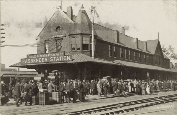 View across railroad tracks towards the depot and platform for the Chicago and Northwestern Railway. The platform is crowded with passengers.