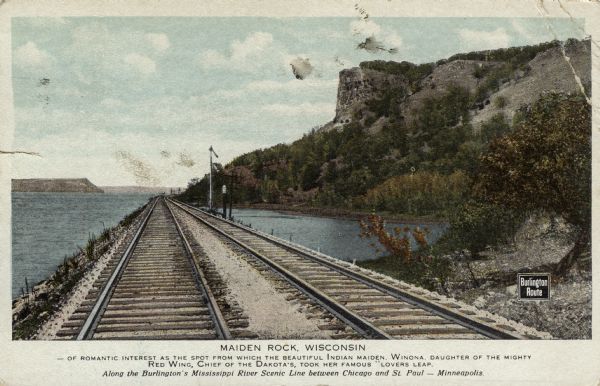 View down railroad tracks along shoreline. Printed sign at right reads: "Burlington Route." Text at bottom reads: "Maiden Rock, Wisconsin — of romantic interest as the spot from which the beautiful Indian maiden, Winona, daughter of the mighty Red Wing, Chief of the Dakotas, took her famous lover's leap. Along the Burlington's Mississippi River Scenic Line between Chicago and St. Paul — Minnesota."