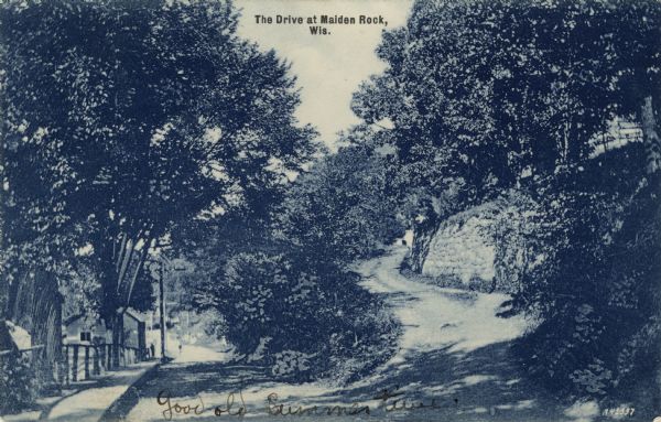 A view of two roads. The road on the left is heading downhill and has a sidewalk along it on the left and buildings in the distance. The other road is winding up a hillside on the right. Caption reads: "The Drive at Maiden Rock, Wis."