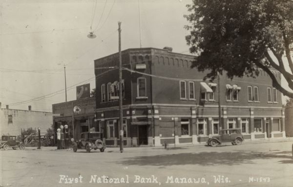 View across intersection towards the corner bank, which is next to a service station. Automobiles are parked at the curb. Caption reads: "First National Bank, Manawa, Wis."