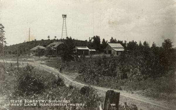 View towards a group of buildings across a road, housing state forestry offices, stables and storage. A watchtower is on a hill. Caption reads: "State Forestry Buildings at Rest Lake, Manitowish Waters."