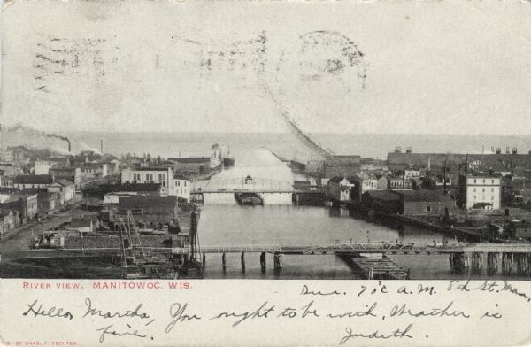 Elevated view over the Manitowoc River where it meets Lake Michigan. Downtown Manitowoc is on both sides, and people are on bridges crossing over the river. Caption reads: "River View, Manitowoc, Wis."