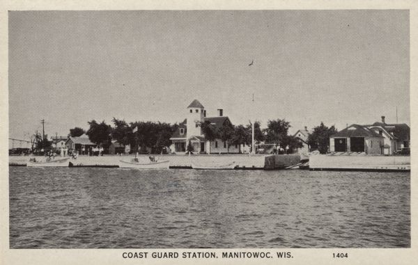 Photographic postcard view across water towards the Coast Guard Station on Lake Michigan. Cutters are moored next to a sea wall. Caption reads: "Coast Guard Station, Manitowoc, Wis."