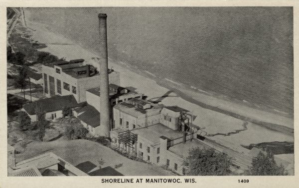 Elevated view of a stretch of beach and a factory with a smokestack along Lake Michigan. Caption reads: "Shoreline at Manitowoc, Wis."