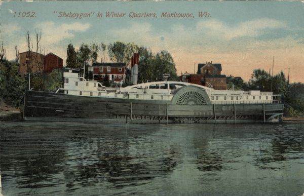 View across water towards a paddle wheel steamer moored for the season. Caption reads: "'Sheboygan' in Winter Quarters, Manitowoc, Wis."