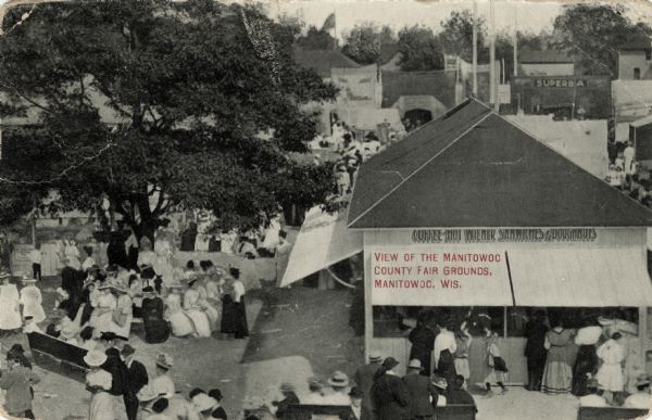 Elevated view of the fairgrounds during the fair. A food vendor is in the foreground and a crowd is gathered there. Caption reads: "View of the Manitowoc County Fair Grounds, Manitowoc, Wis."