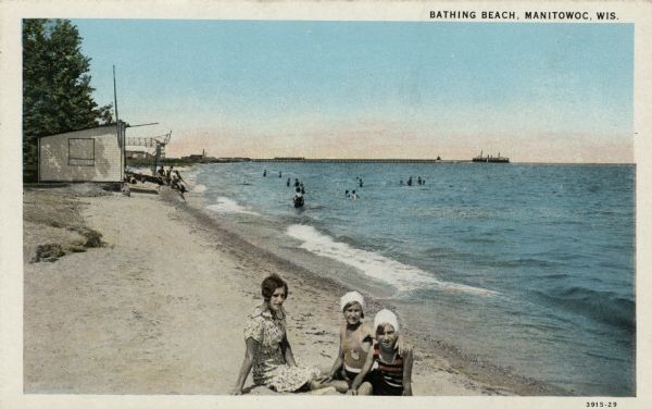 View of a Lake Michigan beach. Three girls are sitting on the sand in the foreground. In the background people are in the water. A steamer is on the horizon. Caption reads: "Bathing Beach, Manitowoc, Wis."