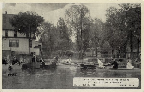 View of Silver Lake Resort (from the lake). Several people are in rowboats, and some of the people have fishing poles. Cottages are among the trees in the background. Text on front reads: "3 1/2 miles west of Manitowoc. M.L. Ouker, Prop."