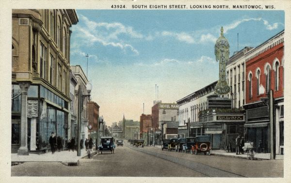 View of the businesses along Eighth Street. The Capitol Theatre and Hotel Manitowoc are on the right. Automobiles are parked along the curbs, and streetcar tracks are running down the center of the street. Caption reads: "South Eighth Street, Looking North, Manitowoc, Wis."