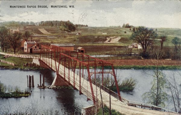 Elevated view of an automobile bridge crossing the Manitowoc River. Rolling hills are in the background. Caption reads: "Manitowoc Rapids Bridge, Manitowoc, Wis."